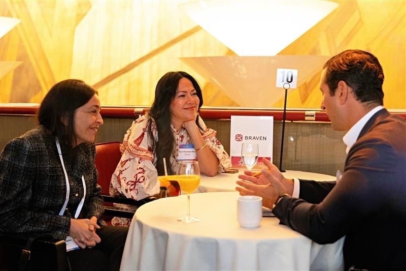 Three professionals engage in a conversation at a Braven-hosted table during a nonprofit networking event, seated with beverages and a table sign marked “10.”