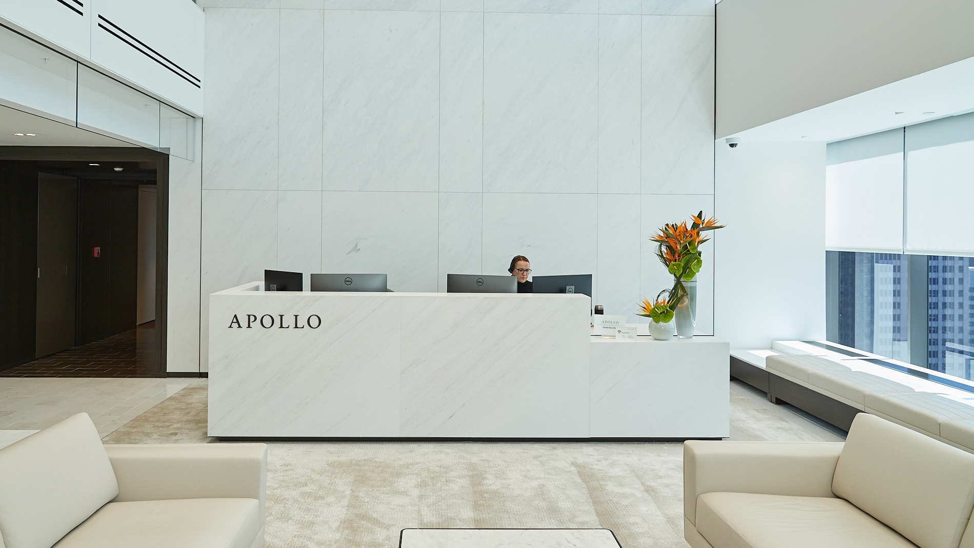 Modern Apollo Global Management reception area with white marble walls, a front desk, seated receptionist, flower arrangement, and city views.