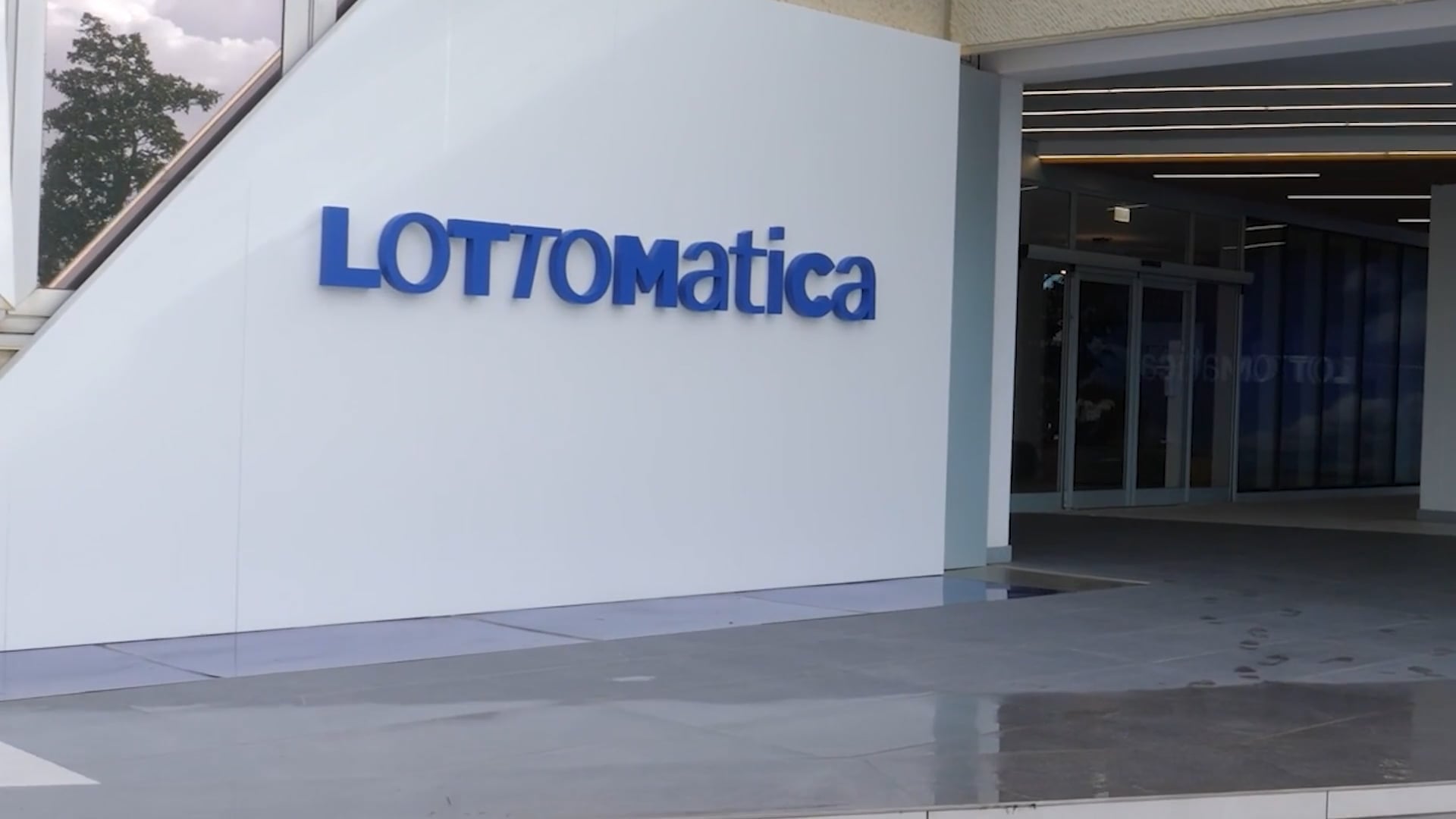 Exterior view of Lottomatica's headquarters entrance, featuring the company’s blue logo on a white wall and glass doors leading into the building.