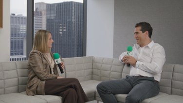 John Cortese and a female interviewer seated on a modern office sofa, discussing private credit trends during a 9@9 video segment with city skyscrapers visible through the window.