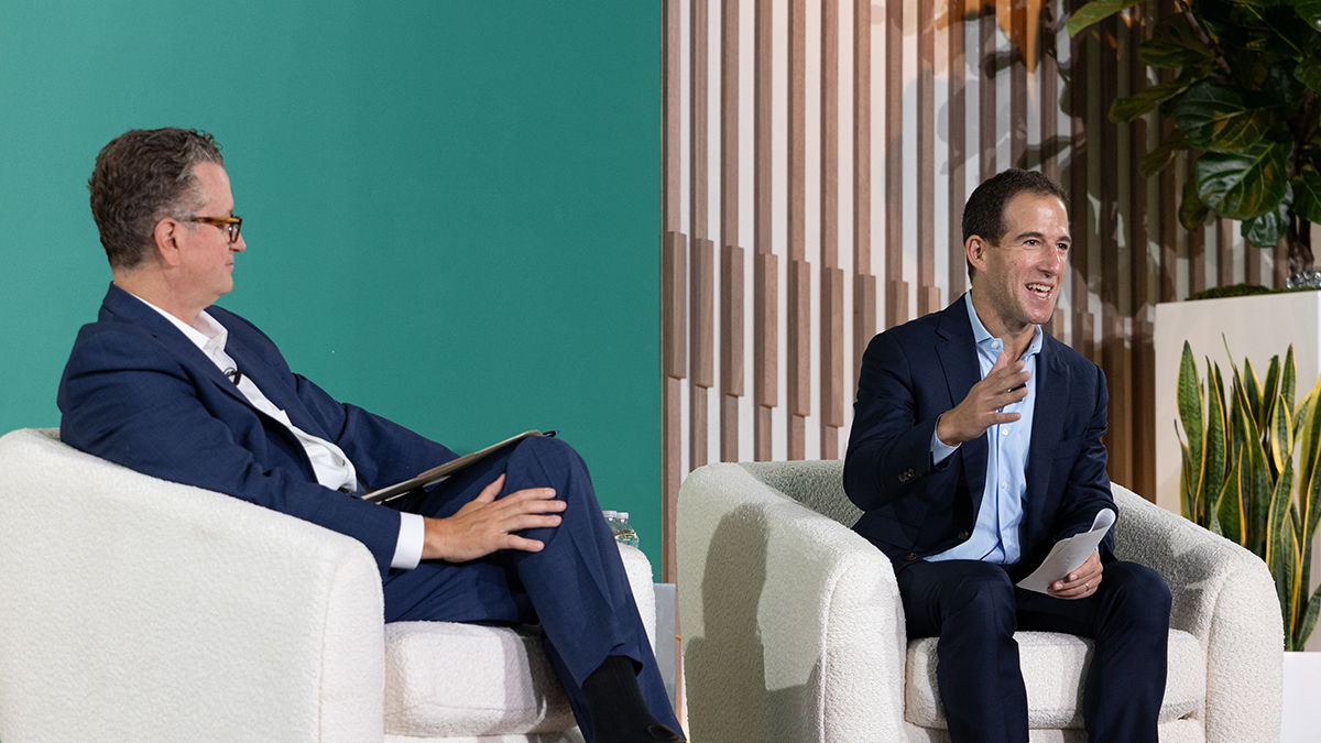 Two executives in navy suits speak during the 2025 Apollo CEO Conference, seated in white armchairs on stage, with modern wood paneling and greenery in the background.